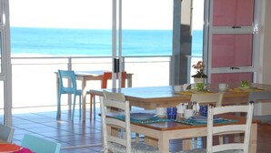 Un coin repas avec vue sur l’océan, comprenant une table et des chaises en bois, une plante en pot et une fenêtre avec des stores roses.