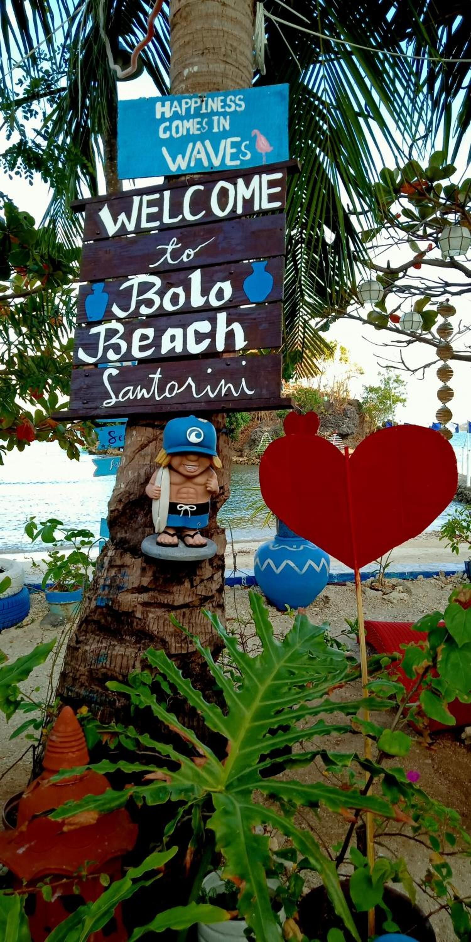 A wooden sign welcoming visitors to Bolo Beach Santorini, featuring a lifeguard figure and a heart-shaped decoration.