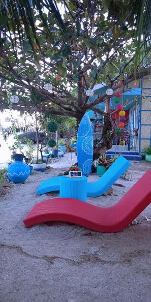 A beachside lounge area with blue and red lounge chairs, a surfboard, and potted plants. - Bolo Beach Santorini (Alaminos)