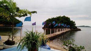 A waterfront view with a wooden dock extending into calm water, surrounded by lush greenery and a row of flags. - Bolo Beach Santorini (Alaminos)
