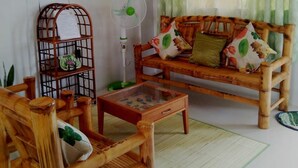 A living room with a wooden sofa, a wooden chair, a glass coffee table, a wall-mounted TV, a standing fan, and a birdcage. - Marie's Sleep Transient House (Alaminos)