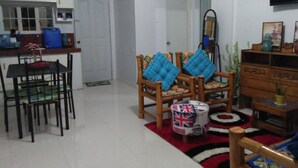A living room with a dining table, two wooden chairs, a sofa with blue cushions, a small wooden side table, and a television mounted on the wall. - Marie's Sleep Transient House (Alaminos)