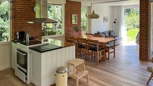 Une cuisine avec un mur en briques, une table à manger en bois et des chaises. Une fenêtre laisse entrer la lumière naturelle et offre une vue sur la verdure à l’extérieur.