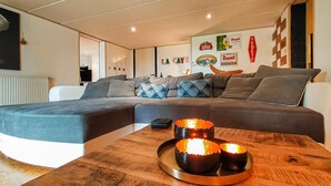 Un salon moderne avec un canapé d’angle, une table basse en bois ornée de bougies et un mur décoré de divers panneaux et affiches.