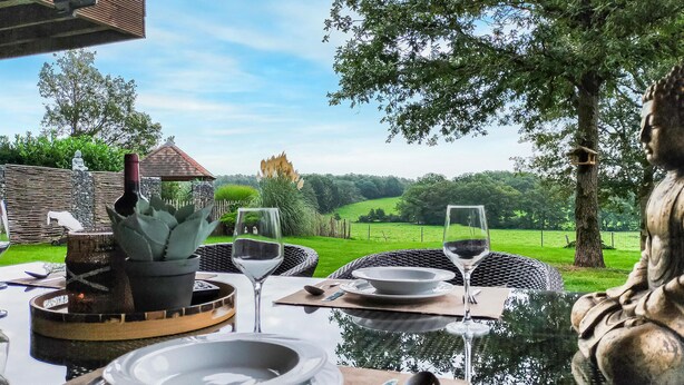 Un espace de restauration en plein air offrant une vue sur un paysage verdoyant, une statue, ainsi qu’une table dressée avec des assiettes, des verres et des couverts.