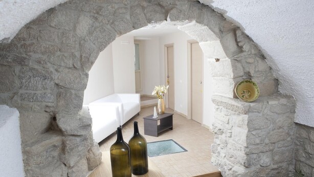 A stone archway leading into a room with a white bed, two bottles, and a small table with a vase of flowers. - B&B Dinastia (Monterosso Almo)