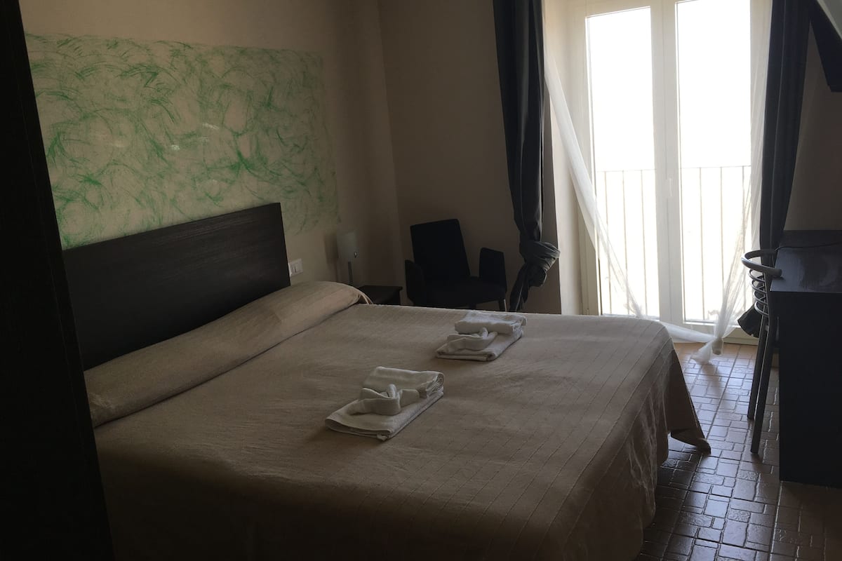 A hotel room with a bed, a chair, a desk, and a balcony with curtains.