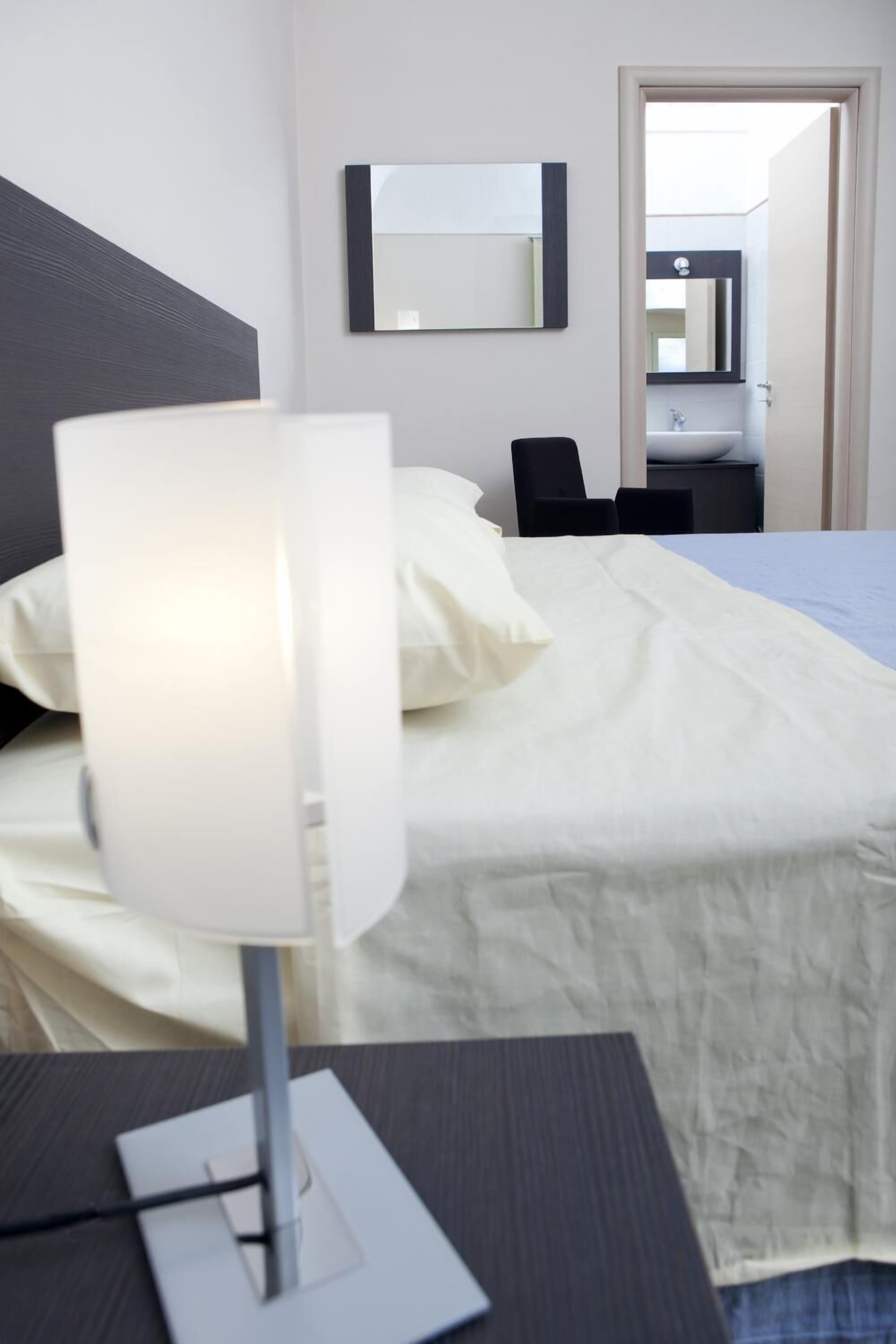 Habitación de hotel con cama, una mesita de noche con lámpara, una silla, un espejo y vistas al baño.