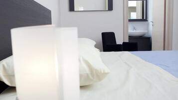 Habitación de hotel con cama, una mesita de noche con lámpara, una silla, un espejo y vistas al baño.