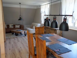 A cozy living room with a sofa, a wooden coffee table, and a dining table with chairs. There are three pendant lights above the table and a hanging light fixture in the corner.