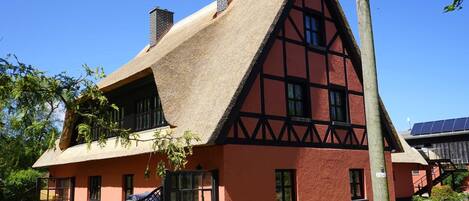 A traditional half-timbered house with a steeply pitched thatched roof, a red facade, and a black gate.