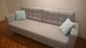 Ein hellgraues Sofa mit gebogener Rückenlehne und zwei hellblauen Kissen, auf einem Holzboden vor einer schlichten Wand platziert.