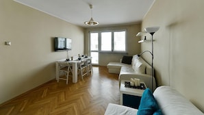 Ein modernes Wohnzimmer mit einer Couch, einem Esstisch mit Stühlen, einem an der Wand befestigten Fernseher und einem großen Fenster, das für natürliches Licht sorgt.