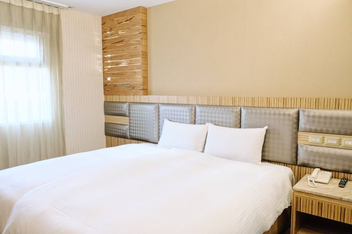 A hotel room with a large bed, a wooden headboard, a bedside table with a phone and remote control, and a window with sheer curtains.