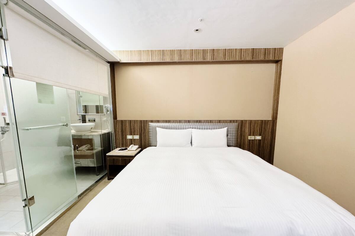 A hotel room with a large bed, a bedside table with a phone, a nightstand, a glass shower enclosure, and a wooden headboard.