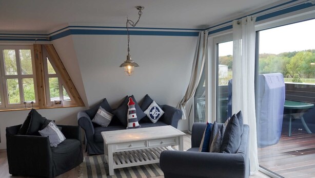 Un salon chaleureux avec un canapé, un fauteuil et une table basse, un luminaire suspendu et une grande fenêtre avec des rideaux.
