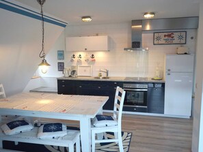 A compact kitchen with white cabinets, a stainless steel oven, and a refrigerator. There is a dining table with chairs and a hanging light fixture.