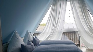 Une chambre avec un plafond en pente, un lit avec une literie bleue et des rideaux en voile.