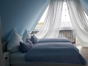 Une chambre avec un plafond en pente, un lit avec une literie bleue et des rideaux en voile.