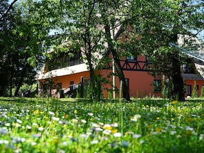 Ein Gebäude mit roter Fassade und dunkler Holzerkerung, umgeben von üppiger Vegetation und einem Feld mit Wildblumen.