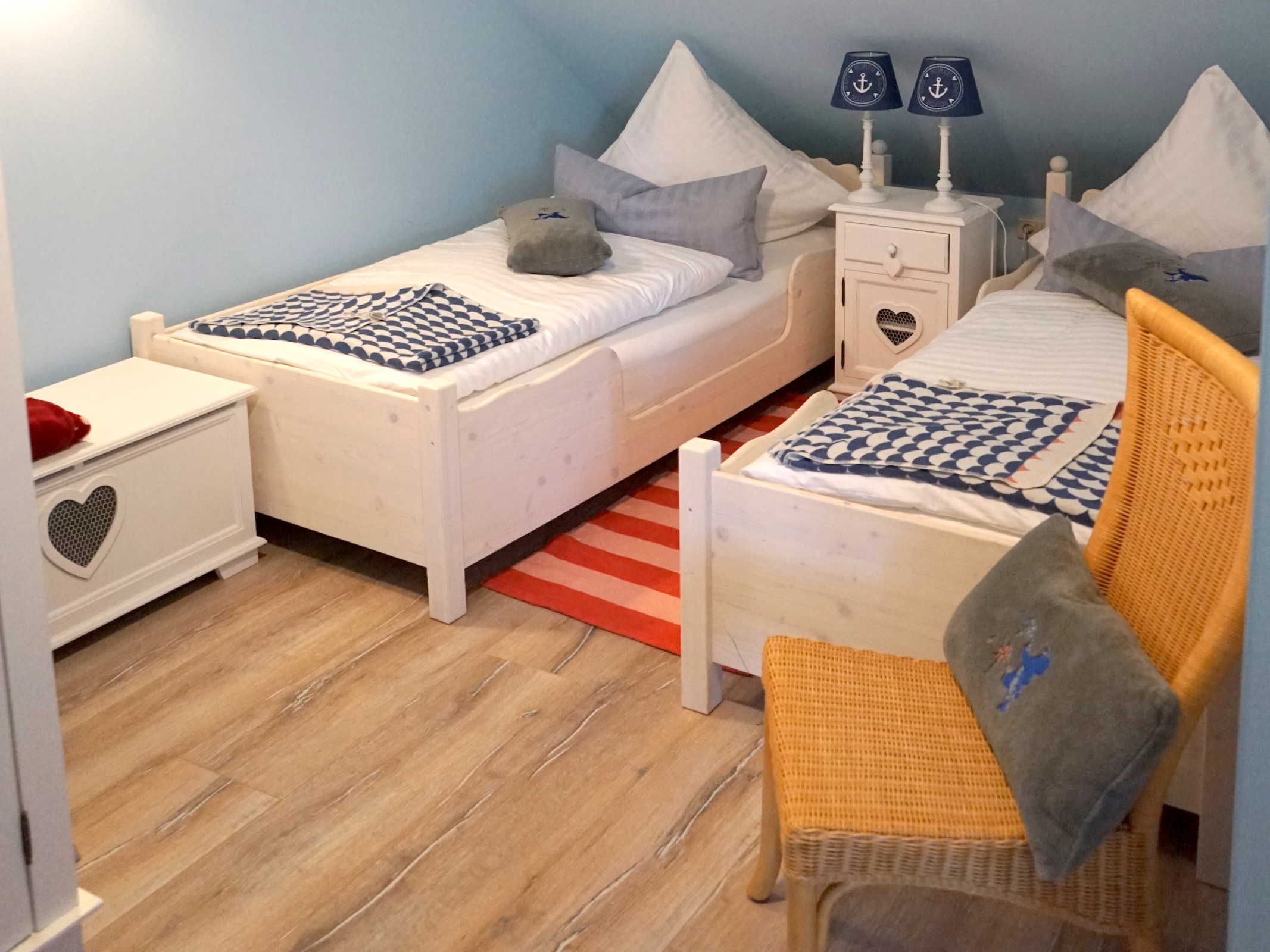 Ein Kinderzimmer mit zwei Einzelbetten, einem kleinen Stuhl, einem Nachttisch mit herzförmiger Schublade und einer Lampe mit Anker-Design.