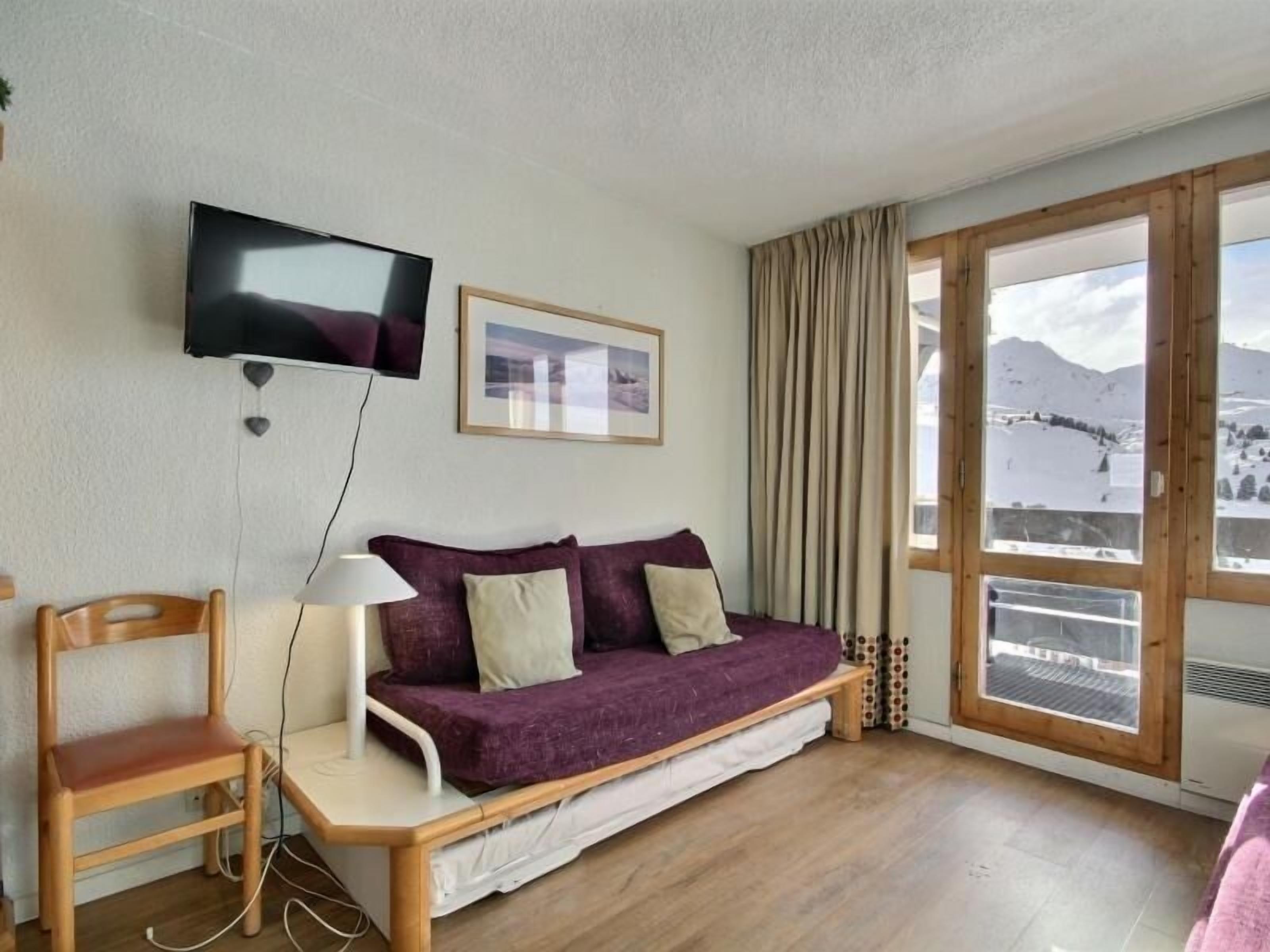 A cozy room with a wooden floor, a purple sofa, a wooden chair, a TV mounted on the wall, and a window with a snowy view.