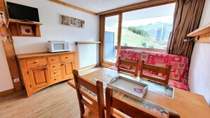 A cozy room with a wooden table, chairs, a red cushioned bench, a television, a microwave, and a window with a view of a green landscape. - Ski-in studio in the heart of Les Menuires, pets allowed (Les Menuires)