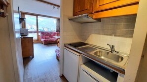 A compact kitchen with a sink, stove, and wooden cabinets. A small seating area with a sofa and a table is visible in the background. - Ski-in studio in the heart of Les Menuires, pets allowed (Les Menuires)