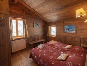 A cozy wooden cabin bedroom with a large bed, two windows, a bench, and a radiator.