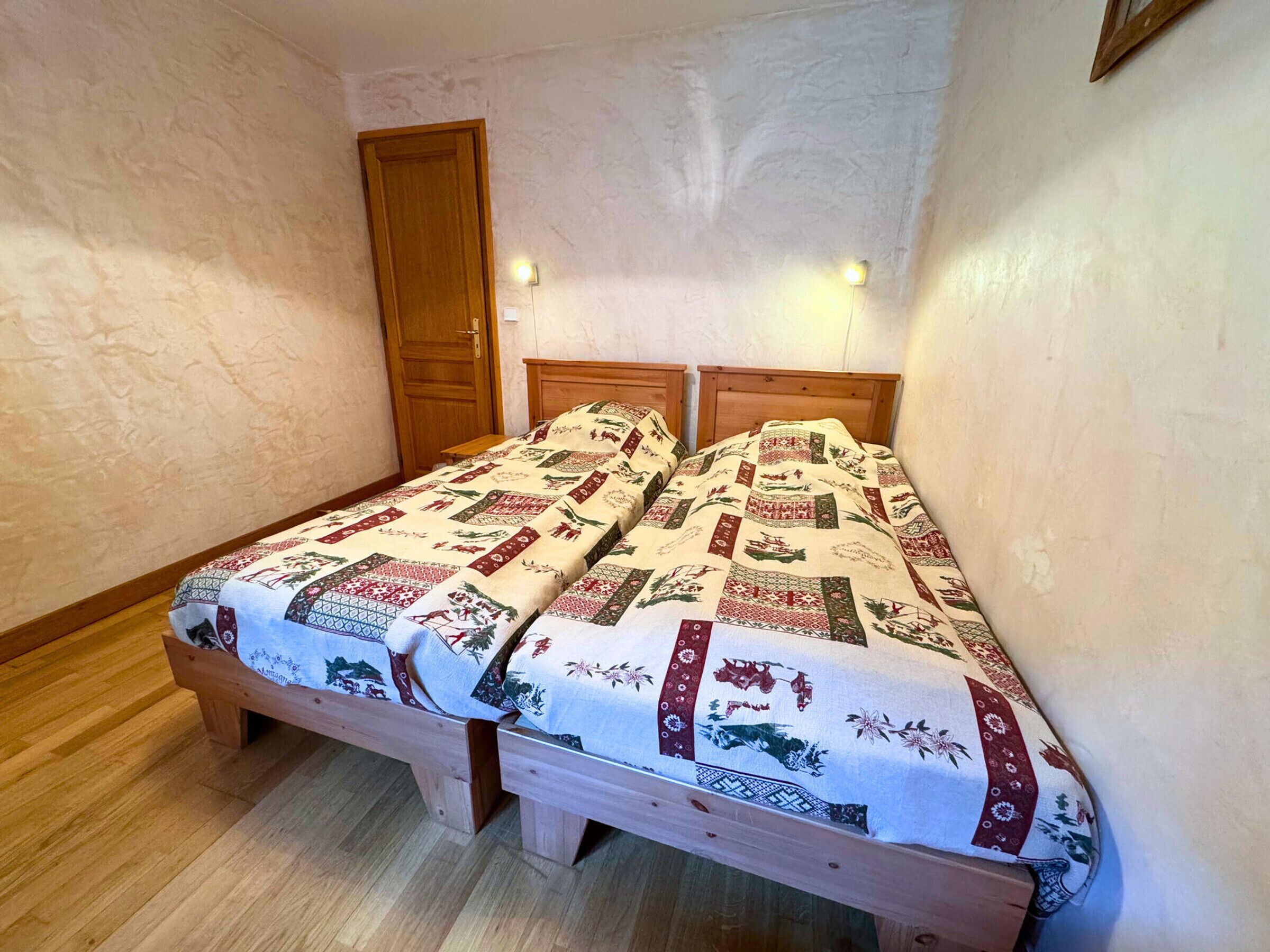 Une petite pièce avec deux lits en bois, chacun recouvert d’une couverture à motifs. On y trouve une porte en bois et un interrupteur sur le mur.