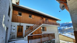 A two-story wooden house with a balcony, a staircase, and a view of snow-covered mountains.