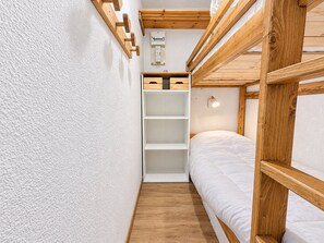 A bunk bed room with a white wall, wooden beams, and a shelf unit.