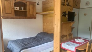 Une petite pièce comprenant un lit, une petite table en bois avec une chaise, un téléviseur et un meuble avec une étagère.