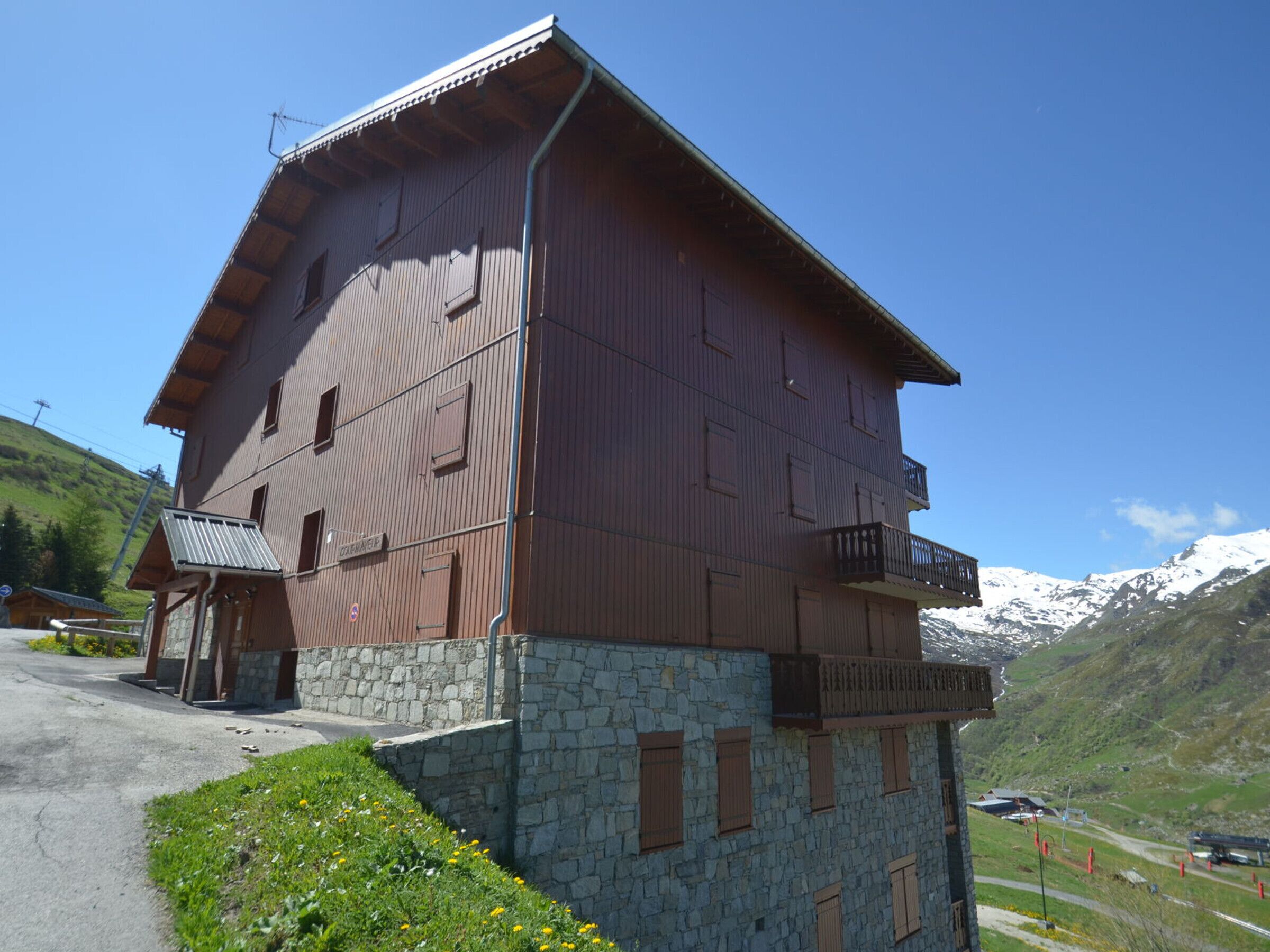 A two-story wooden building with stone foundations, a balcony on the upper floor, and a clear view of snow-capped mountains in the background.