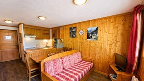 Een compacte kamer met een houten bank, een kleine televisie, een eettafel en een kitchenette.
