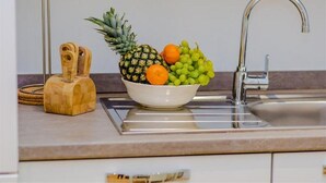 Una encimera de cocina con un frutero que contiene piña, naranjas y uvas, una tabla de cortar de madera y un grifo moderno.