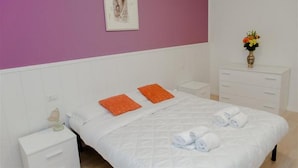 Una cama bien tendida con ropa blanca y almohadas naranjas, una mesita de noche con una lámpara y un florero con flores sobre un tocador.