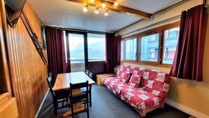 Una habitación acogedora con una escalera de madera, una mesa de comedor con sillas, un sofá con un cubrecolchón estampado en rojo y blanco, y ventanas grandes con cortinas.
