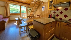 Een gezellig vakantiehuisje met houten inrichting, een eethoek met tafel en stoelen, een keuken met fornuis en kastjes, en een raam met uitzicht op groen.