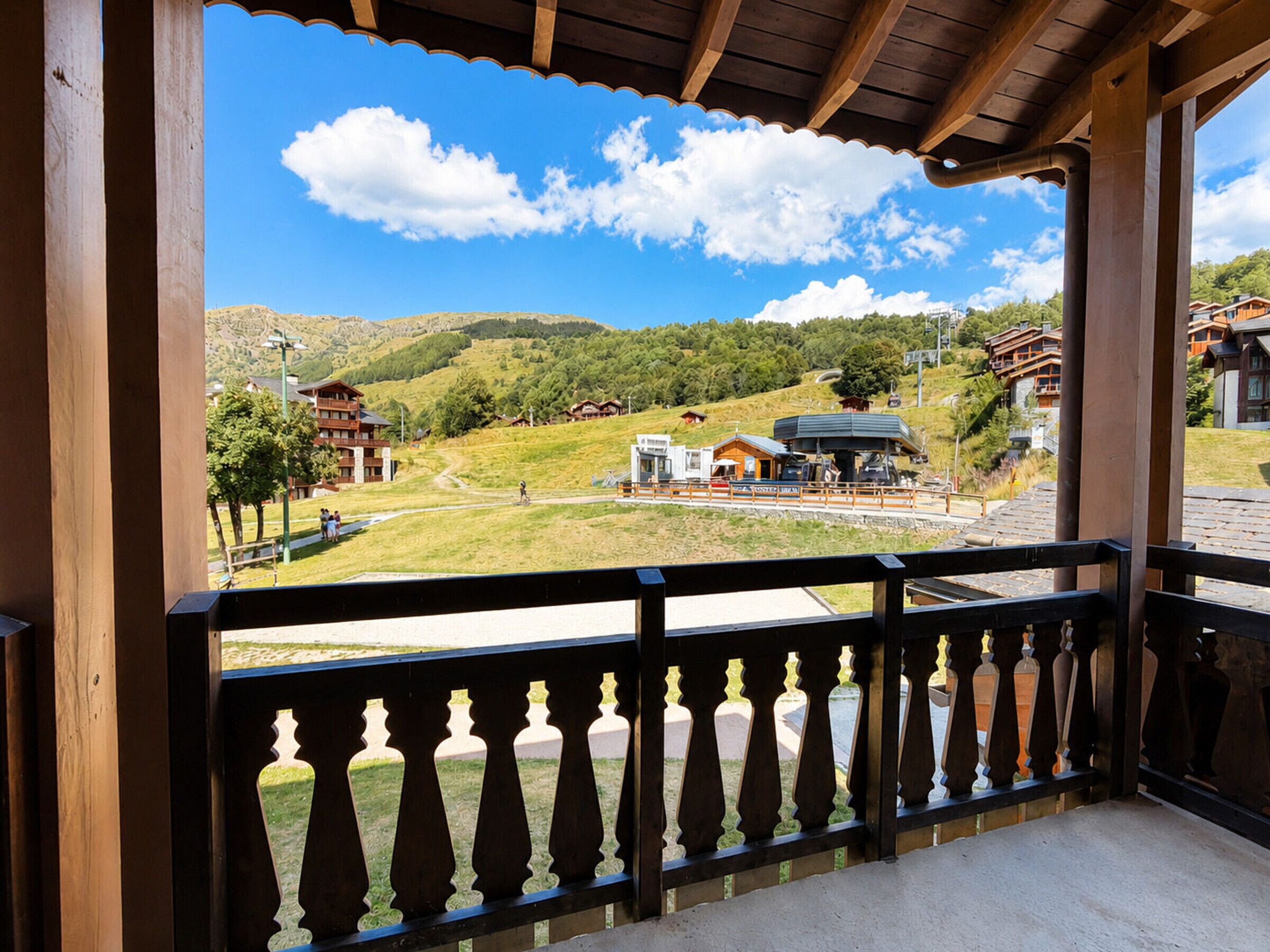 A wooden balcony with a view of a mountainous landscape, including buildings and a ski lift.