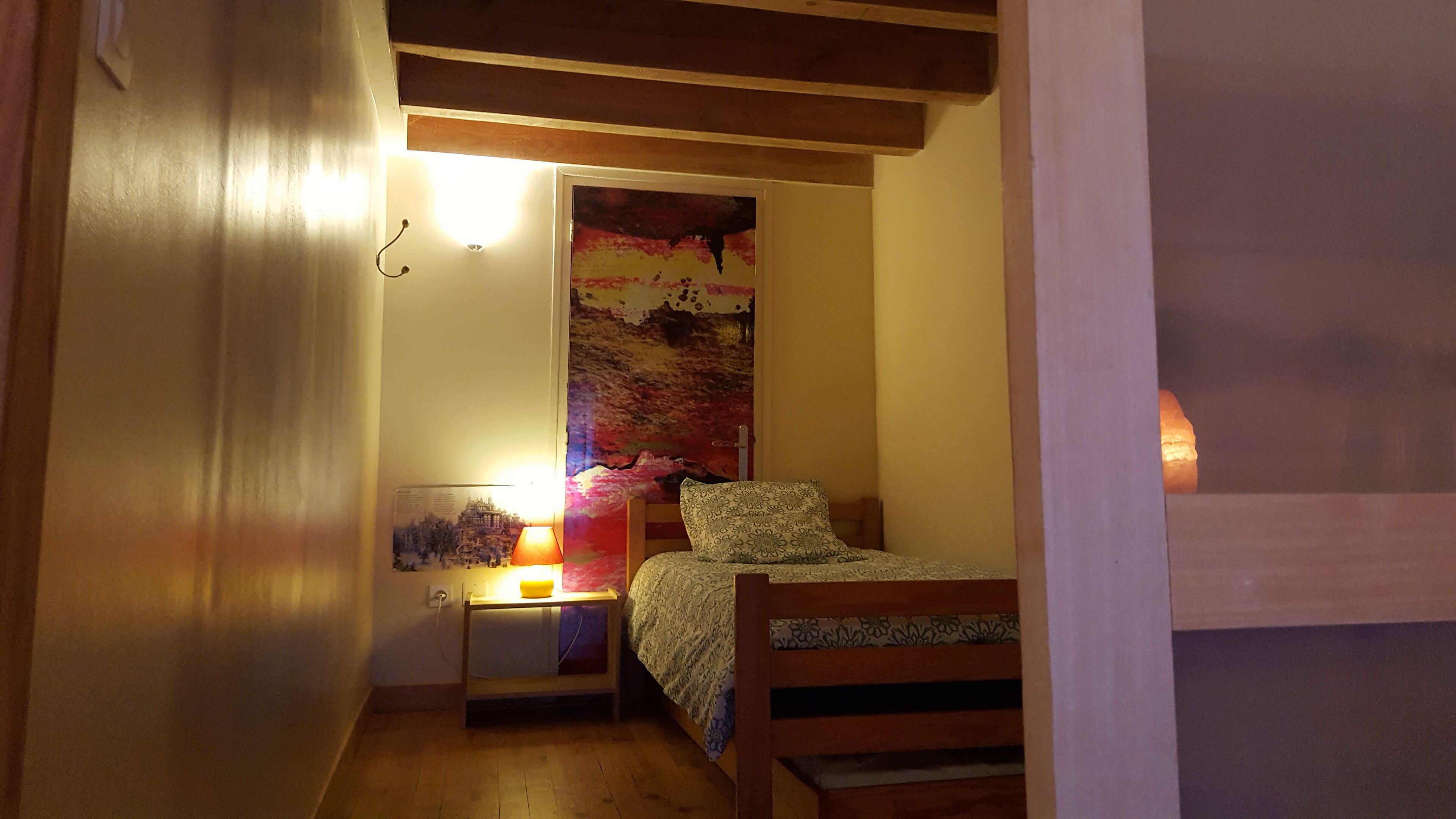 A small bedroom with a bed, a nightstand with a lamp, and a wall-mounted shelf.