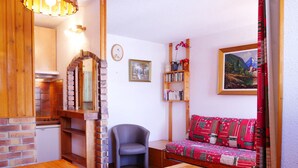 A cozy living room with a wooden dining table, a sofa with a patterned blanket, a bookshelf, and a painting on the wall.