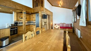 A cozy cabin-style room with a wooden dining table, chairs, a kitchen area with a refrigerator, and a loft space with a bed.