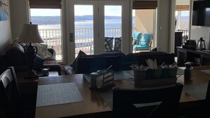 Een woonkamer met een eettafel, stoelen, een bank en een plafondventilator. Er zijn grote ramen met uitzicht op het water en de heuvels.