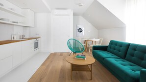Una cocina moderna con armarios blancos y encimera de madera, un sofá verde azulado y una zona de comedor con mesa y sillas blancas.
