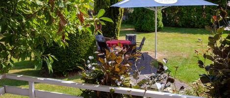 A patio area with a table and chairs, surrounded by lush greenery and a white fence.