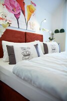 Una cama bien hecha con ropa blanca y almohadas decorativas, un cabecero tapizado en rojo y un cuadro floral en la pared.