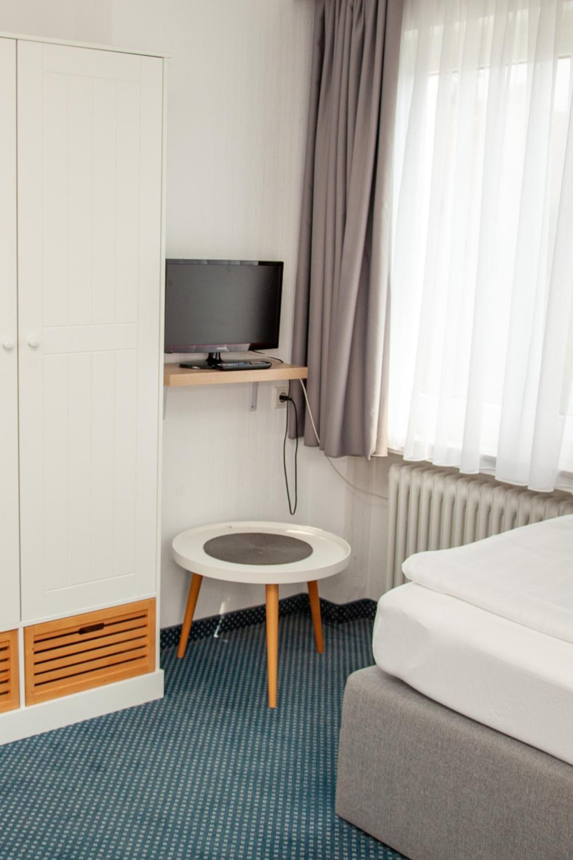 A hotel room with a bed, a TV on a shelf, a round table, and a wardrobe.