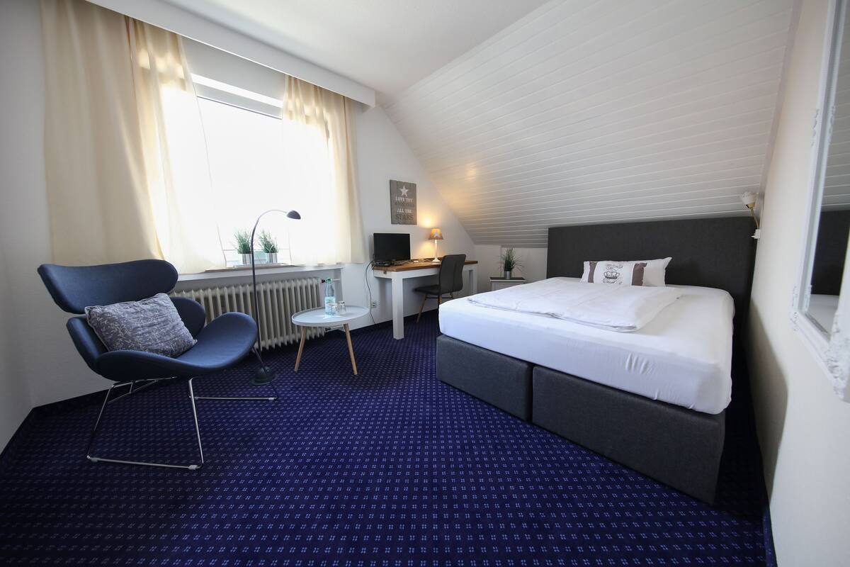 Une chambre d’hôtel avec un lit, un bureau, une chaise, un radiateur et une fenêtre avec des rideaux.