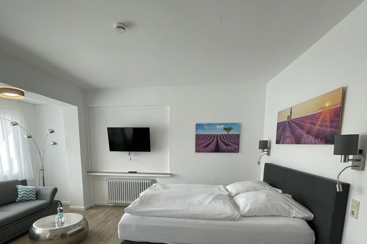 A modern hotel room with a bed, a TV, a sofa, and artwork on the walls.